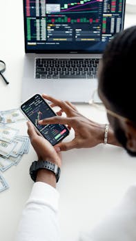 A person trading stocks using a smartphone, showcasing financial data on mobile and laptop screens.