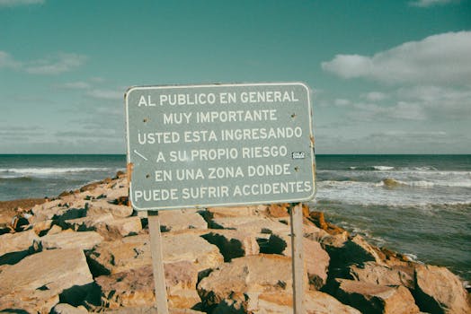 Spanish warning sign by rocky coast with crashing waves under clear skies.